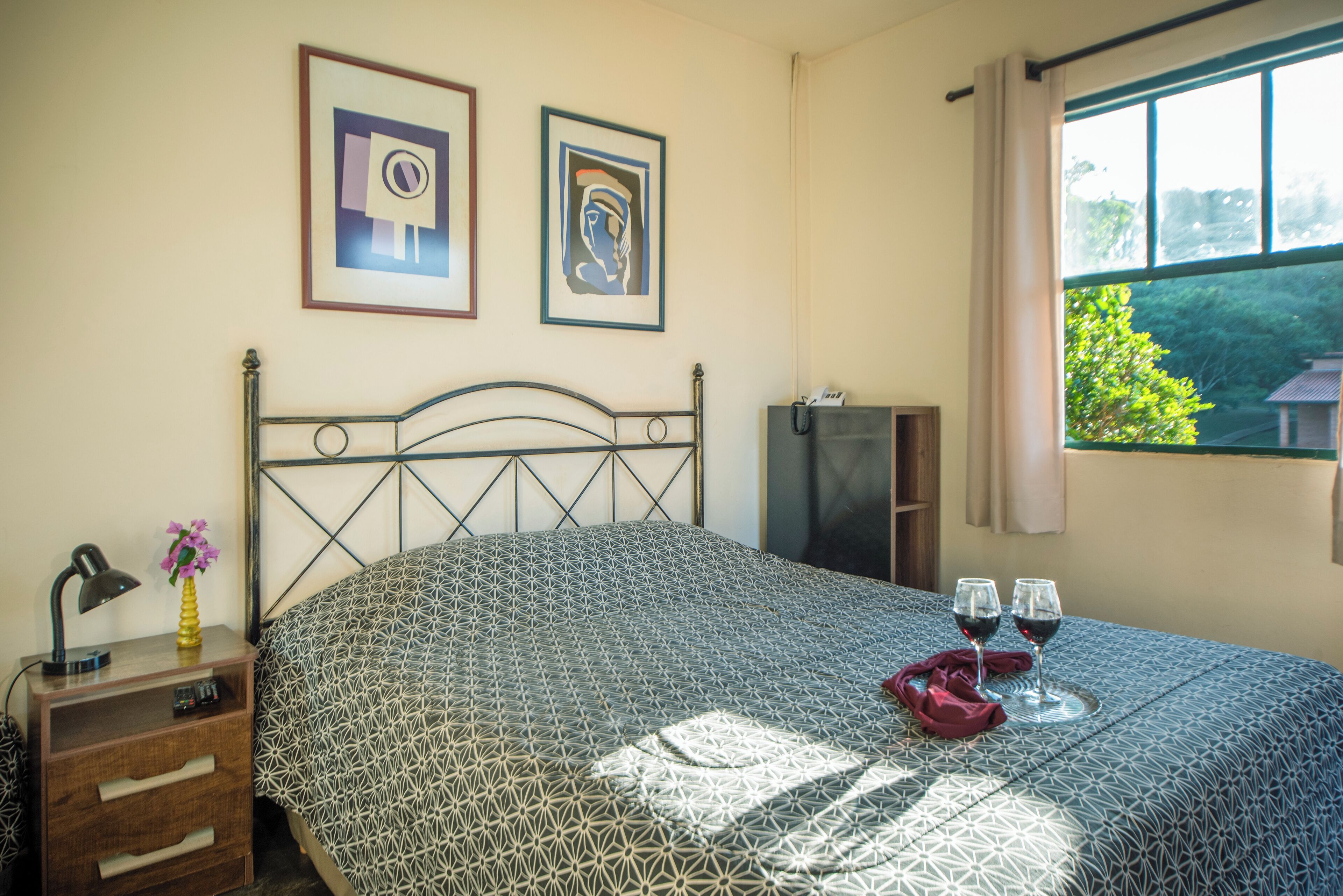 A bedroom with a patterned bedspread, a nightstand with a lamp, a vase with flowers, and two framed pictures on the wall.