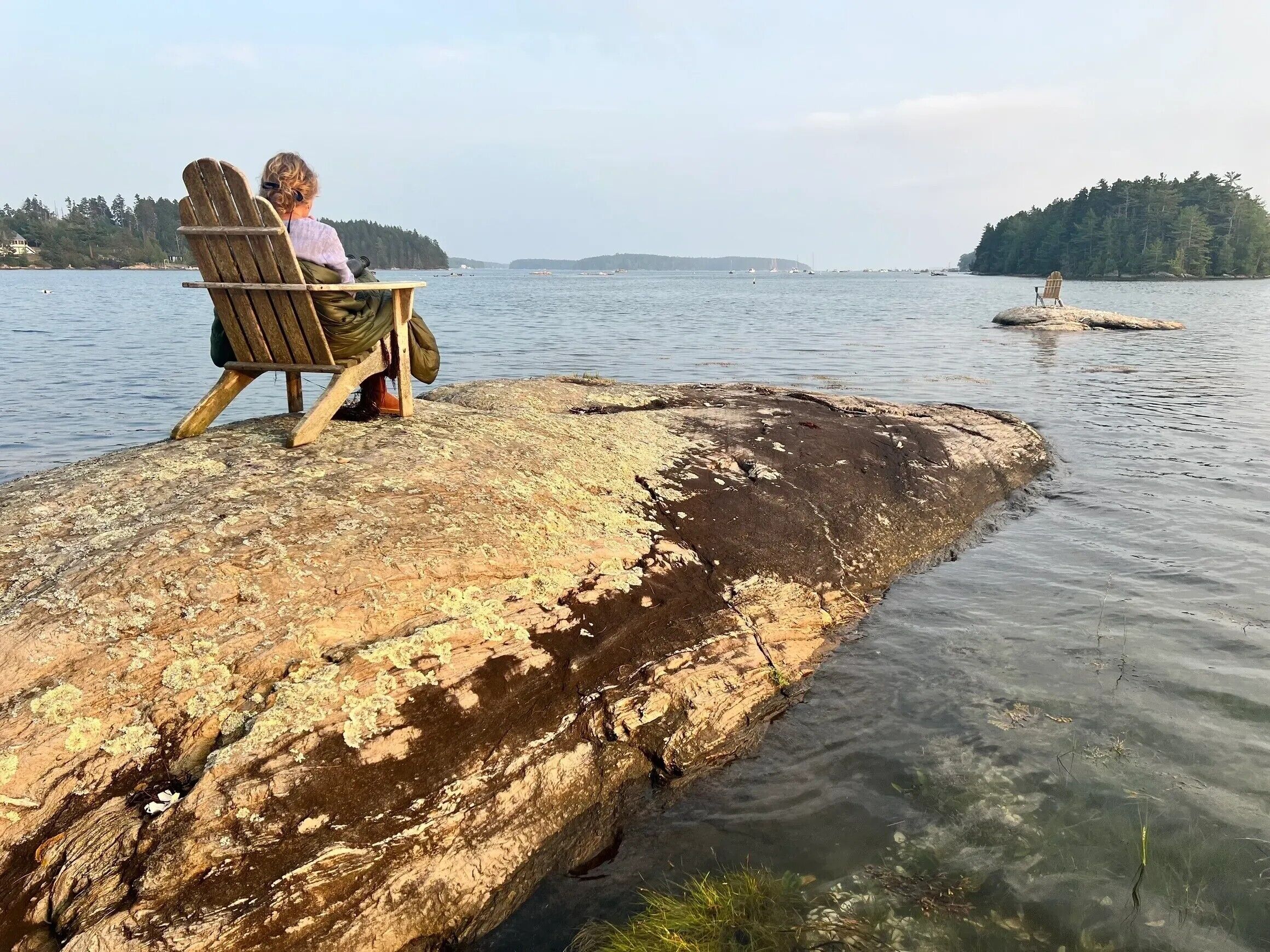 A wooden chair on a rocky outcrop by a calm lake with distant boats and a forested island.
