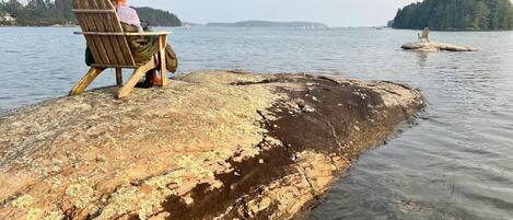 A wooden chair on a rocky outcrop by a calm lake with distant boats and a forested island.