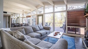 Een ruime woonkamer met een grote grijze hoekbank, een salontafel en een eethoek met stoelen en een kroonluchter.