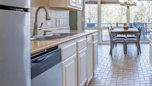Een moderne keuken met witte kasten, een roestvrijstalen koelkast en een vaatwasser. Er is een eethoek met een tafel en stoelen, en grote ramen die voor natuurlijk licht zorgen.