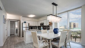 A modern kitchen with white cabinetry, a stainless steel refrigerator, and a dining area with wicker chairs and a white table. - Pelican Beach Terrace 307 * 3 bedroom * Salt Water Vacations (Destin)