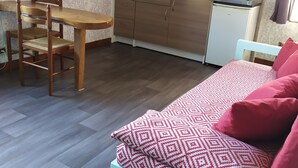 Una habitación pequeña con cama, una mesa de comedor de madera con sillas, una kitchenette y un mueble blanco pequeño.