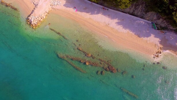 An aerial view of a beach with clear turquoise waters, a sandy shore, and a rocky outcrop.