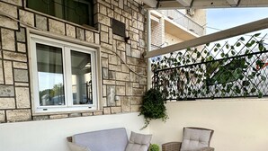 A covered outdoor seating area with wicker furniture, a potted plant, and a window with a view of the street.