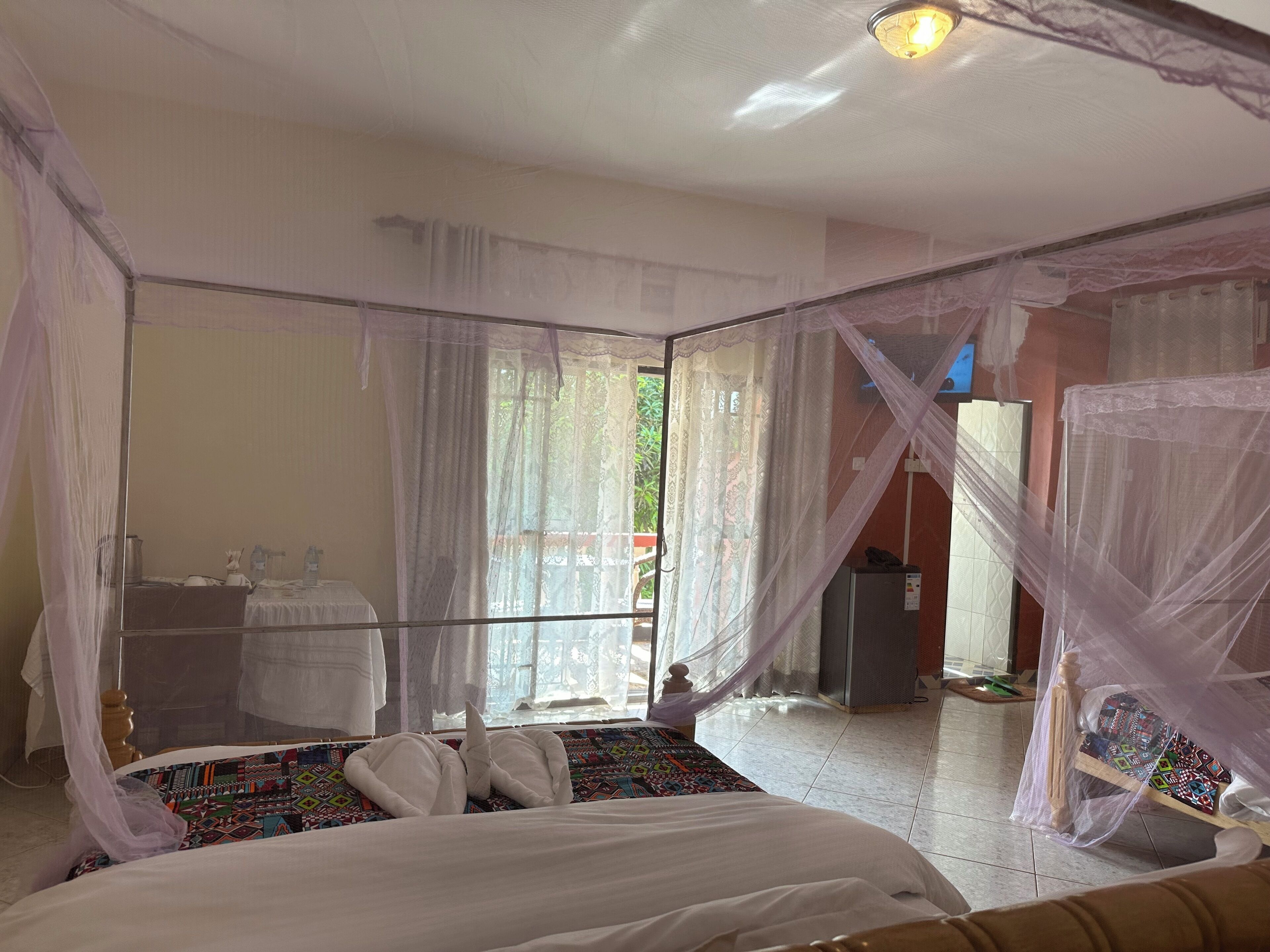 A bedroom with a bed, a bedside table, a mirror, and a window with sheer curtains.