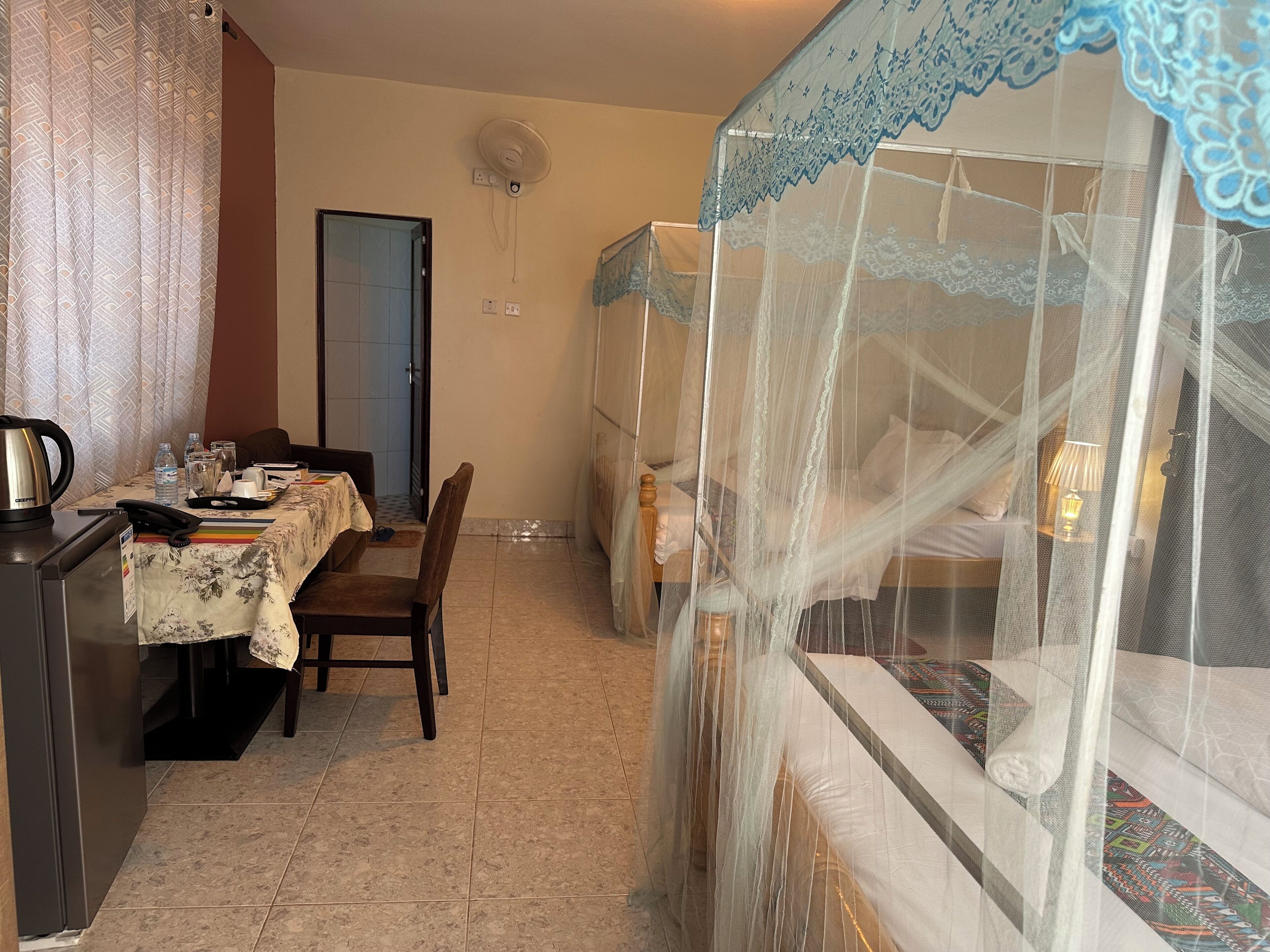 Une chambre d’hôtel comprenant un coin repas, un lit doté d’une moustiquaire, un ventilateur fixé au mur et une porte donnant sur une autre pièce.