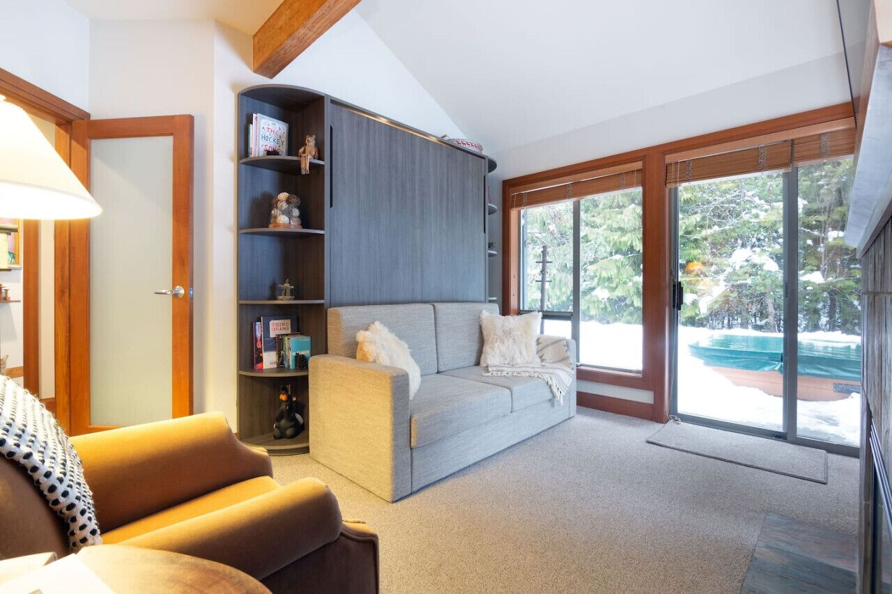 A cozy living room with a grey sofa, a wooden bookshelf, and a large sliding glass door leading to a balcony.