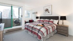 Een slaapkamer met een bed, nachtkastje, bureau en raam met uitzicht op gebouwen.