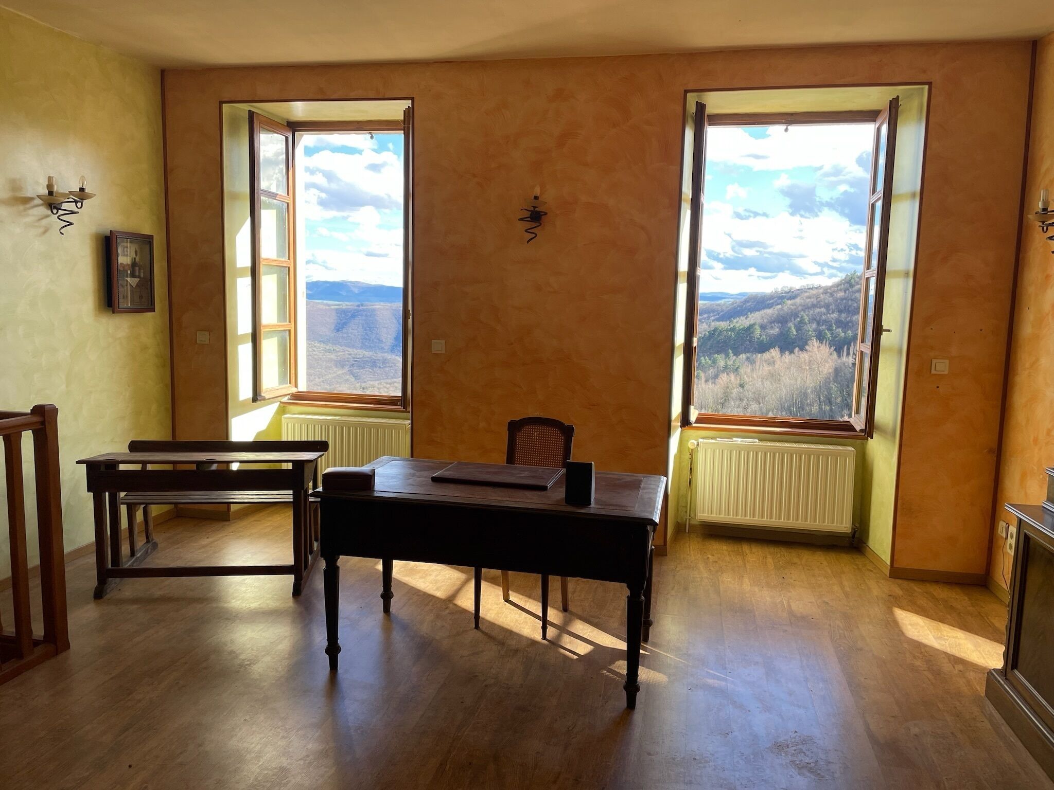 Habitació amb dues finestres grans que donen vistes a un paisatge muntanyós. Al centre de l'habitació hi ha una taula de fusta i una cadira.