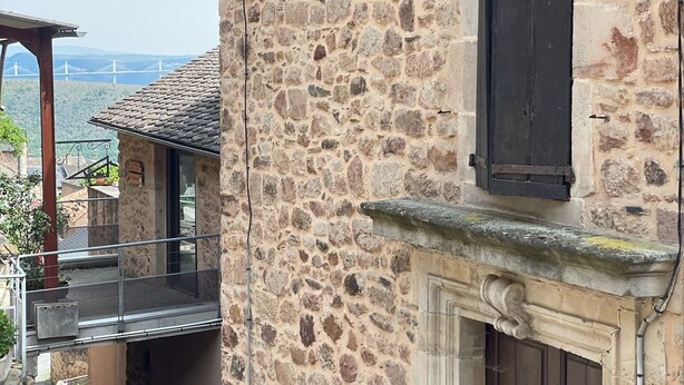 Un bâtiment en pierre doté d’un balcon, d’une cheminée et offrant une vue sur un pont en arrière-plan.