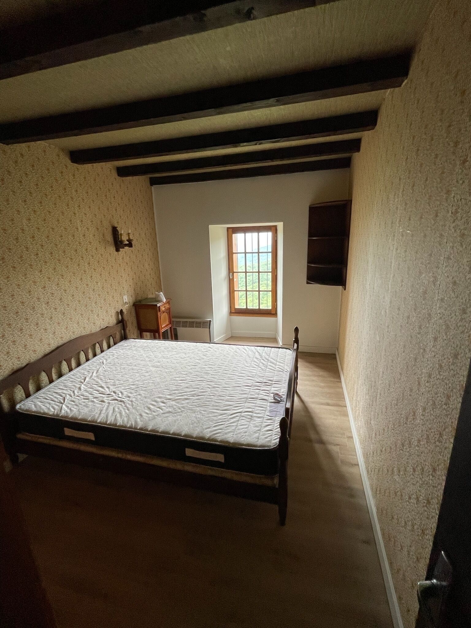 Een slaapkamer met een houten bed, een klein nachtkastje van hout, een raam met uitzicht en een schuine plafond met zichtbare balken.