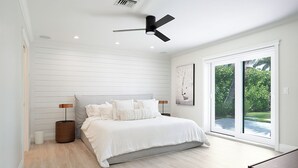 Quarto moderno com cama grande, ventilador de teto e porta de vidro deslizante para um jardim.