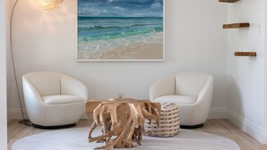 Quarto moderno com uma mesa redonda, duas poltronas brancas, uma mesa de centro de madeira, um quadro com tema praiano e um abajur.