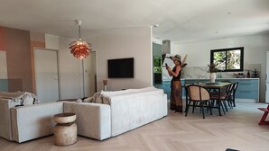 Una sala de estar moderna con un sofá, un comedor con sillas, una cocina al fondo y una persona de pie junto a la ventana.