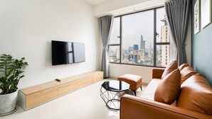 A modern living room with a large window, a flat-screen TV, a sofa, a coffee table, and a potted plant. - Cen Oasis Apart - CenRiver Side (Ho Chi Minh City)
