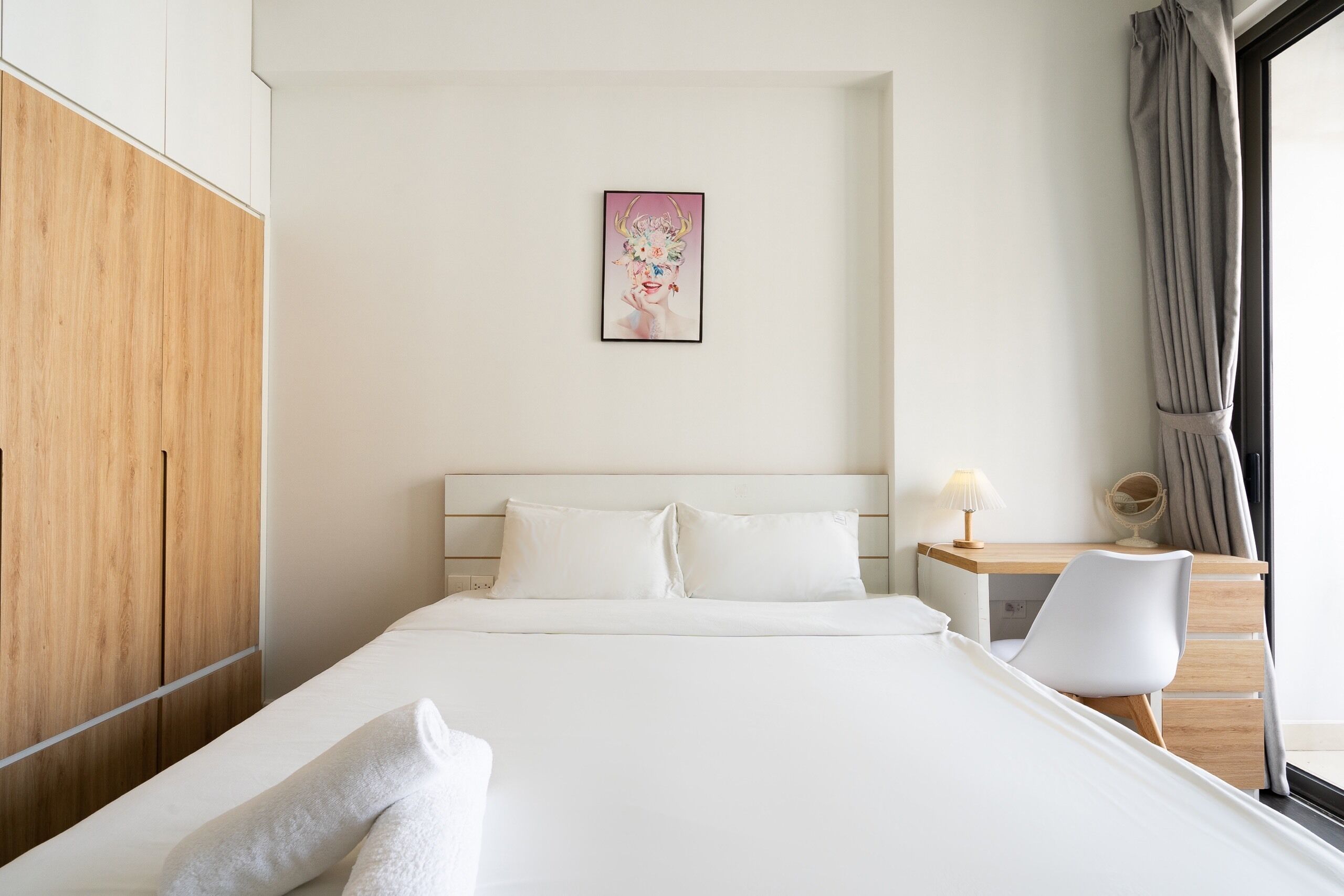 A neatly made bed with white linens, a wooden wardrobe, a desk with a lamp, and a framed picture on the wall.