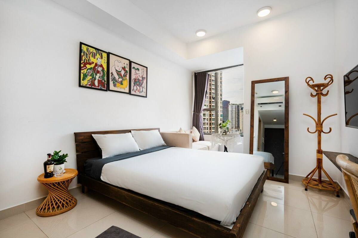 A modern bedroom with a large bed, a bedside table with a bottle and a plant, a mirror, and a view of tall buildings through a window.