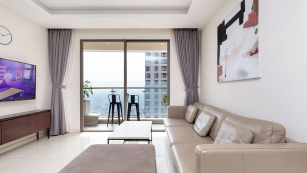 A modern living room with a sofa, a coffee table, and a TV on a wall. There is a balcony with a view of buildings. - Cen Oasis Apart - CenRiver Side (Ho Chi Minh City)