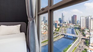 A hotel room with a large window overlooking a cityscape, including a river and modern buildings. - Cen Oasis Apart - CenRiver Side (Ho Chi Minh City)
