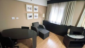 A hotel room with a round table, two armchairs, a sofa, a TV on a stand, and a window with curtains. - Studio Living Mercure Liberdade (São Paulo)