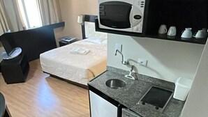 A compact kitchenette with a microwave, sink, and small refrigerator. There is a bed with a white comforter and a chair. The room has wooden flooring and a window with curtains. - Studio Living Mercure Liberdade (São Paulo)