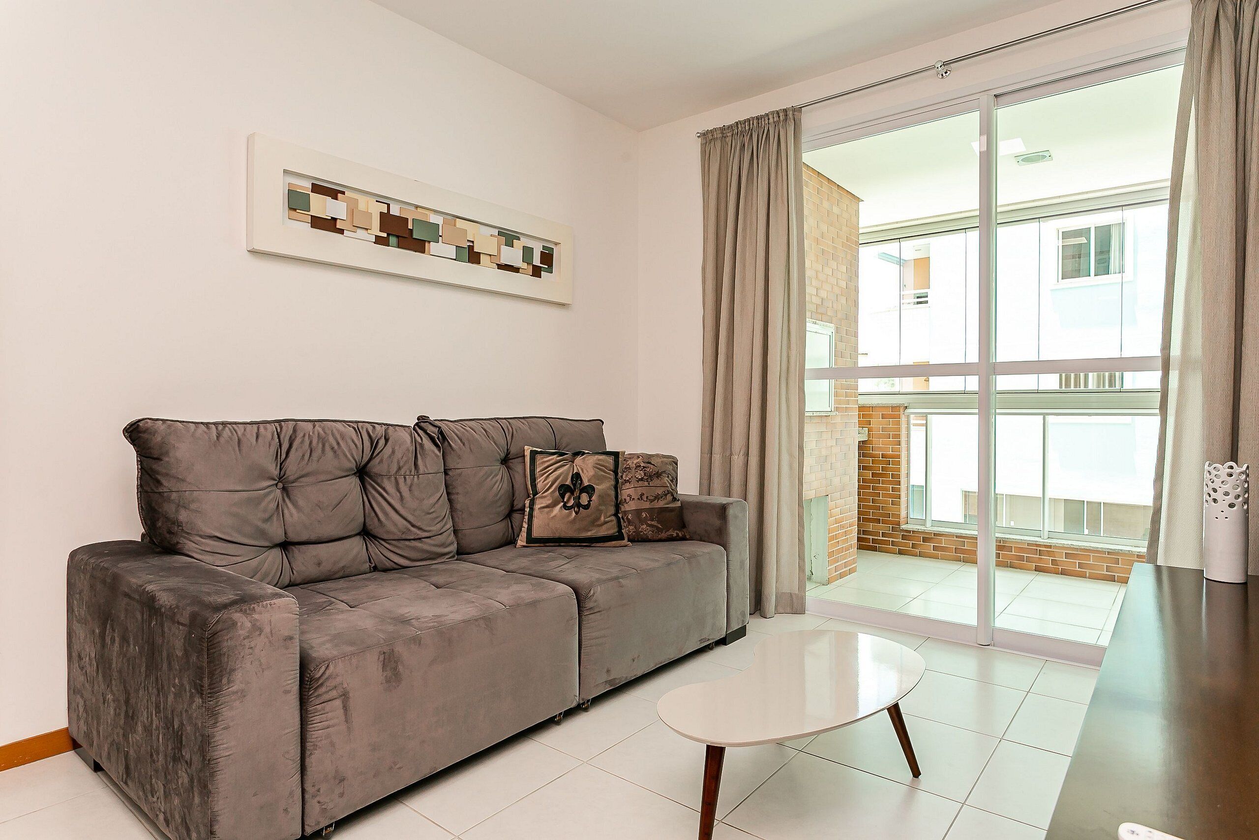 A modern living room with a grey sofa, a white coffee table, and a large sliding glass door leading to a balcony.