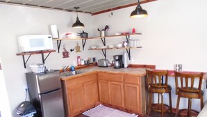 Une cuisine compacte équipée d’un réfrigérateur, d’un four à micro-ondes et de meubles en bois. On y trouve deux chaises en bois autour d’une petite table, et une poubelle est visible.