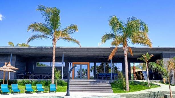A modern building with a pool area, featuring blue lounge chairs and palm trees.