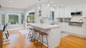 A modern kitchen with white cabinets, a large island with stools, a built-in oven, and a view of greenery outside. - Family Beach House! Remodeled, Walk to Beach + Yard + Deck & Swim Spa (Huntington Beach)