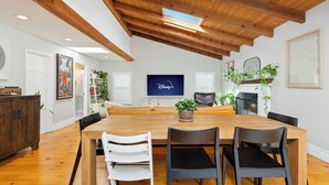 A dining area with a wooden table, four chairs, a fireplace, and a television displaying Disney+. - Family Beach House! Remodeled, Walk to Beach + Yard + Deck & Swim Spa (Huntington Beach)