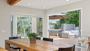 A spacious dining area with a wooden table and chairs, a potted plant, and a view of the outdoor patio with a sofa and umbrella. - Family Beach House! Remodeled, Walk to Beach + Yard + Deck & Swim Spa (Huntington Beach)