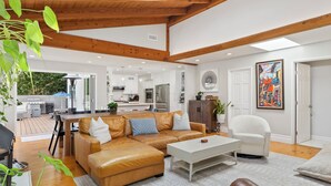 A modern living room with a leather sofa, a white coffee table, and a dining area with a wooden table and chairs. - Family Beach House! Remodeled, Walk to Beach + Yard + Deck & Swim Spa (Huntington Beach)