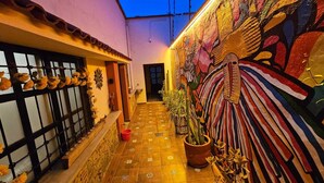 Un pasillo estrecho con suelo de baldosas, un muro decorativo con un mural colorido, y plantas en macetas a los lados.