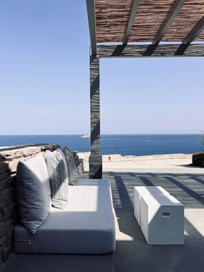 A modern outdoor seating area with a view of the sea, featuring a sofa and a bench under a wooden pergola. - Cave Suite (Paros)