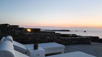En udendørs siddeplads med stenvægge, hvide puder og et bord, med udsigt til havet i solnedgang.