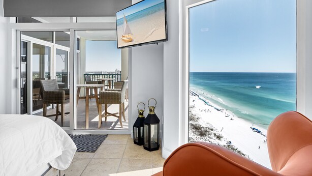 Une chambre d’hôtel dotée d’une grande fenêtre donnant sur une plage, d’un lit, d’une télévision et d’un coin repas avec des chaises et une table.