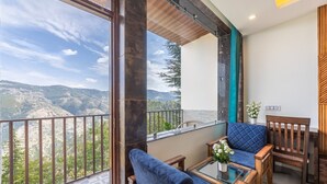 A balcony with a view of mountains, a blue sofa, a wooden chair, and a glass table with a potted plant. - Manaw Valley Resort (Shimla)