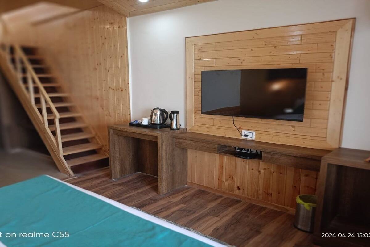 Ein Zimmer mit einer Holztreppe, einem an einer Holzwand montierten Fernseher, einem kleinen Tisch mit Wasserkocher und einem Bett mit türkisblau gestreifter Bettdecke.