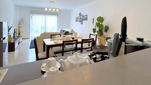A modern living room with a dining area, featuring a large window, a dining table set for four, and a kitchen area with a stove and sink. - E&D  Apartment With Private Garden, Quiet and Secure Ideal For Families (Antananarivo)