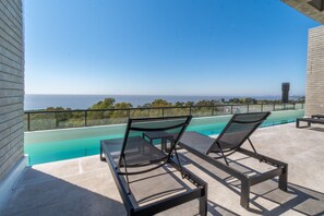 Ein Poolbereich auf dem Dach mit zwei Liegestühlen und einem kleinen Tisch, mit Blick auf einen klaren, blauen Himmel und eine entfernte Meereslandschaft.