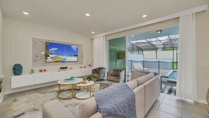 Una sala de estar moderna con un televisor grande, asientos cómodos, un ventanal que da a un balcón y una zona de piscina.