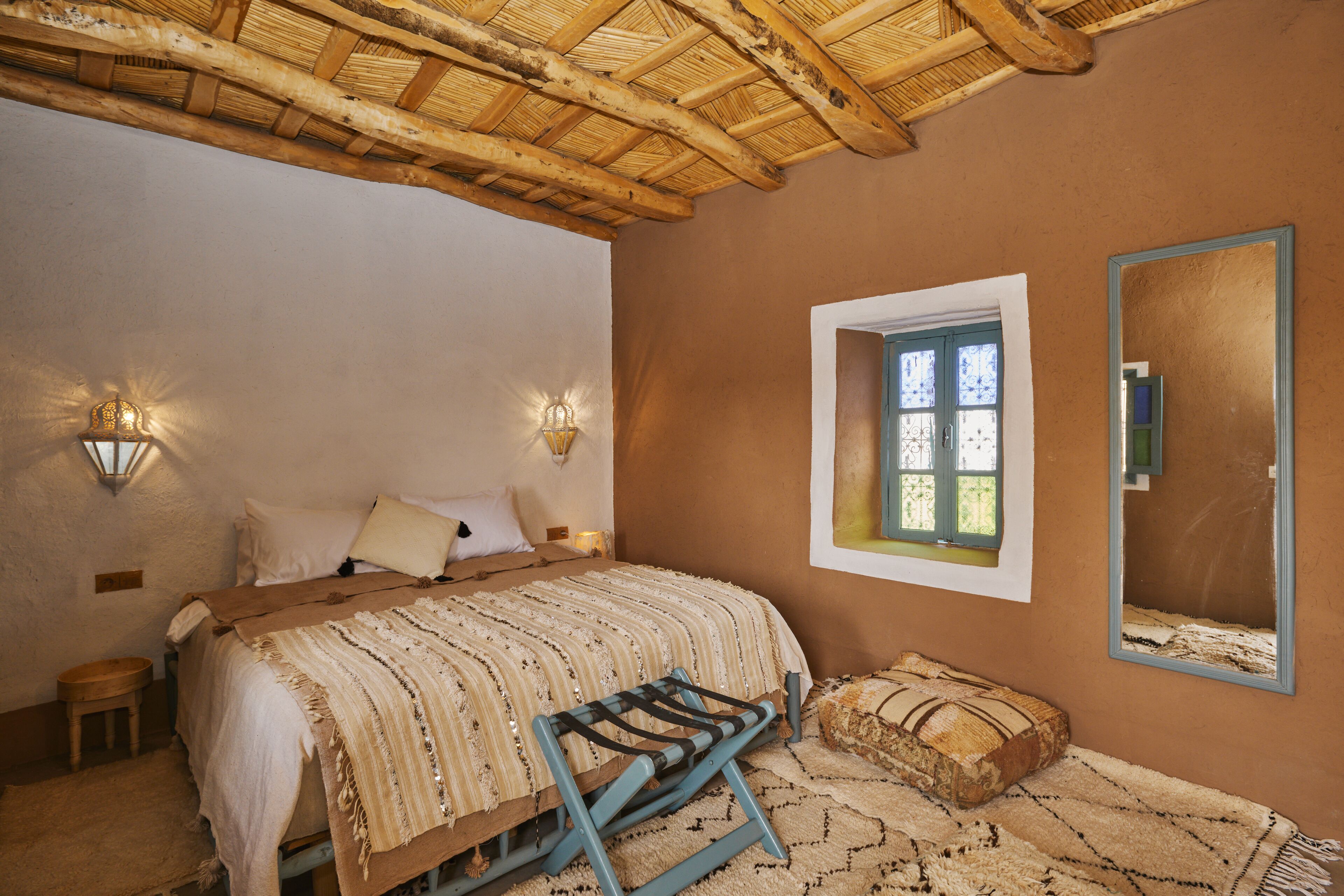 Quarto com cama, mesa de cabeceira, banqueta, janela e espelho.