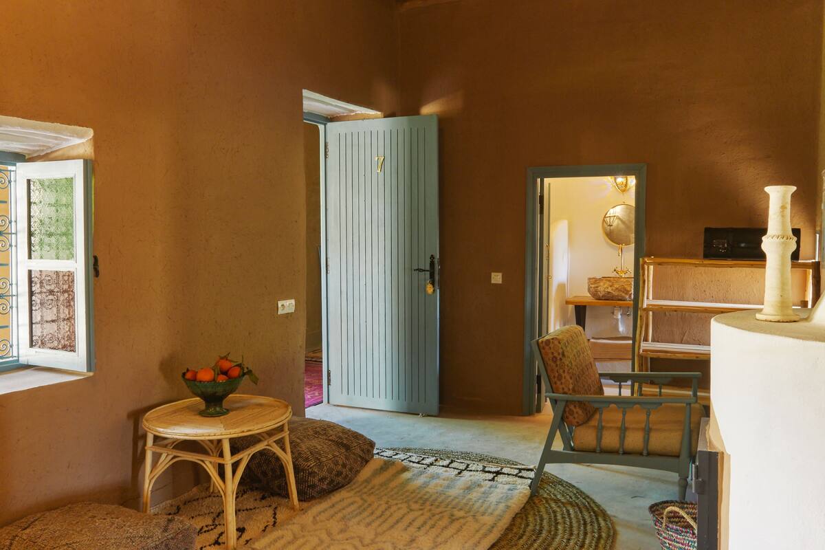 A cozy room with a wicker chair, a small table with a bowl of fruit, a blue door, and a patterned rug.