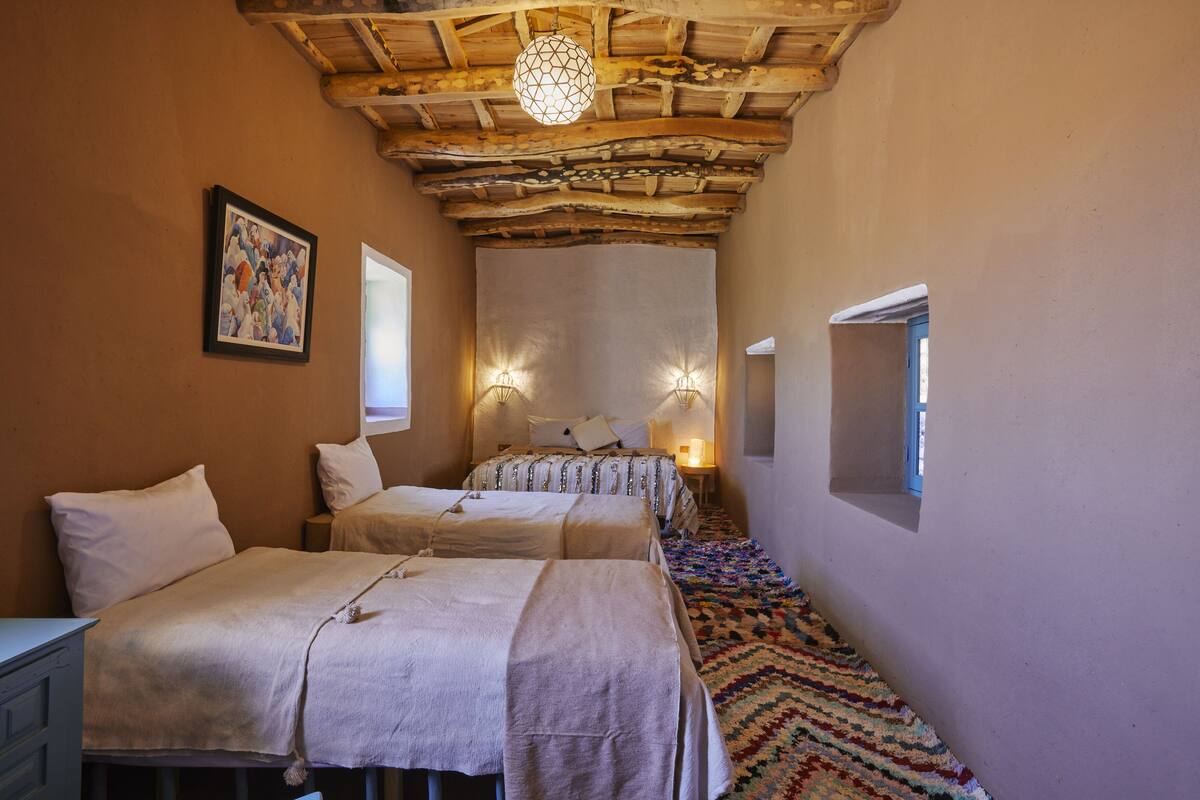 A room with two beds, a wooden ceiling, a patterned carpet, and a painting on the wall.