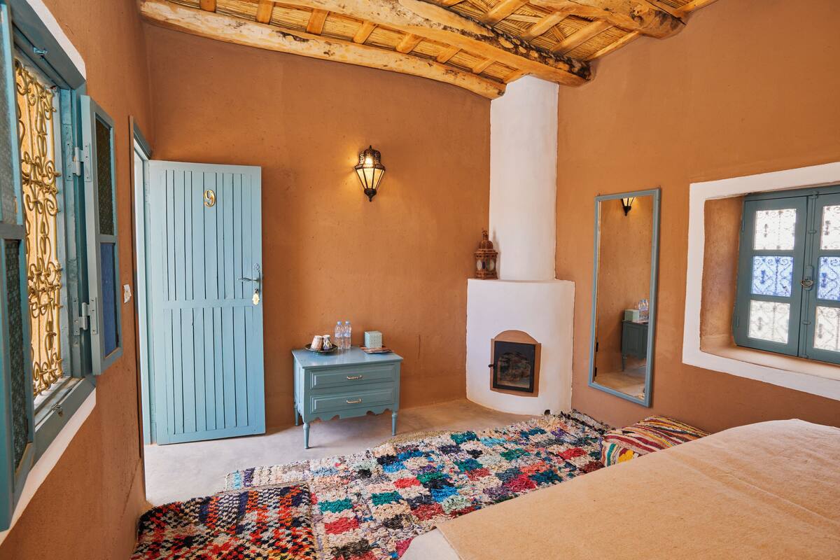 A room with a blue door, a small dresser, a colorful rug, a fireplace, and a large mirror.
