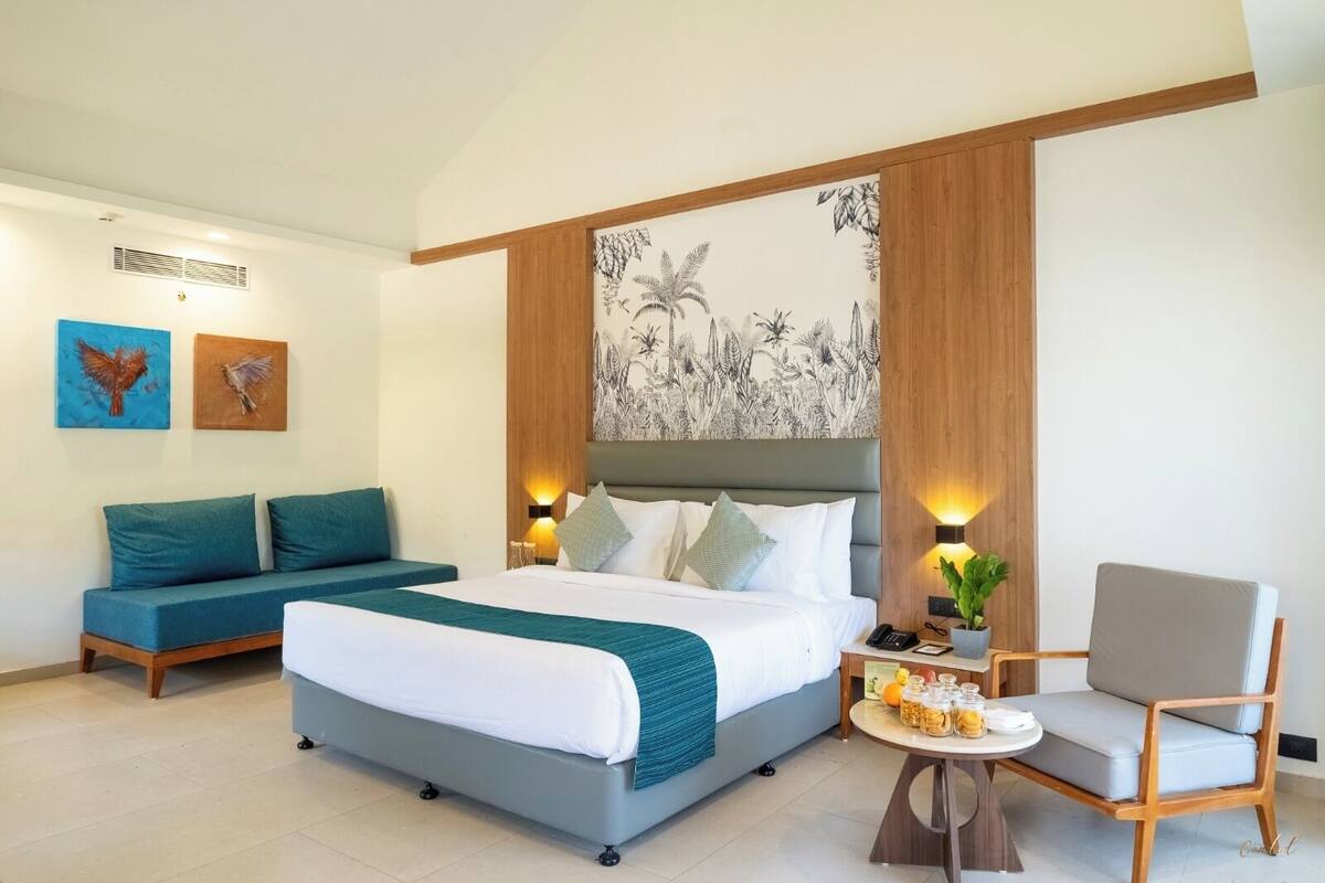 A hotel room with a bed, a sofa, a chair, and a small table with a plant and a phone.