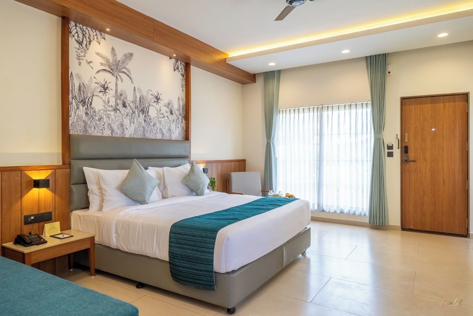 Une chambre d’hôtel moderne avec un grand lit, un bureau avec une lampe, un ventilateur de plafond et une grande fenêtre avec des rideaux.
