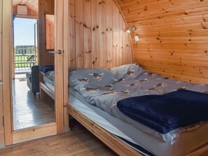 A wooden cabin with a bed featuring a grey blanket with animal prints, a wooden ceiling, and a view of a deck outside.