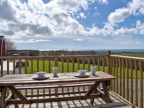 Um deck de madeira com uma mesa posta para dois, com vista para um campo gramado e colinas ao longe.